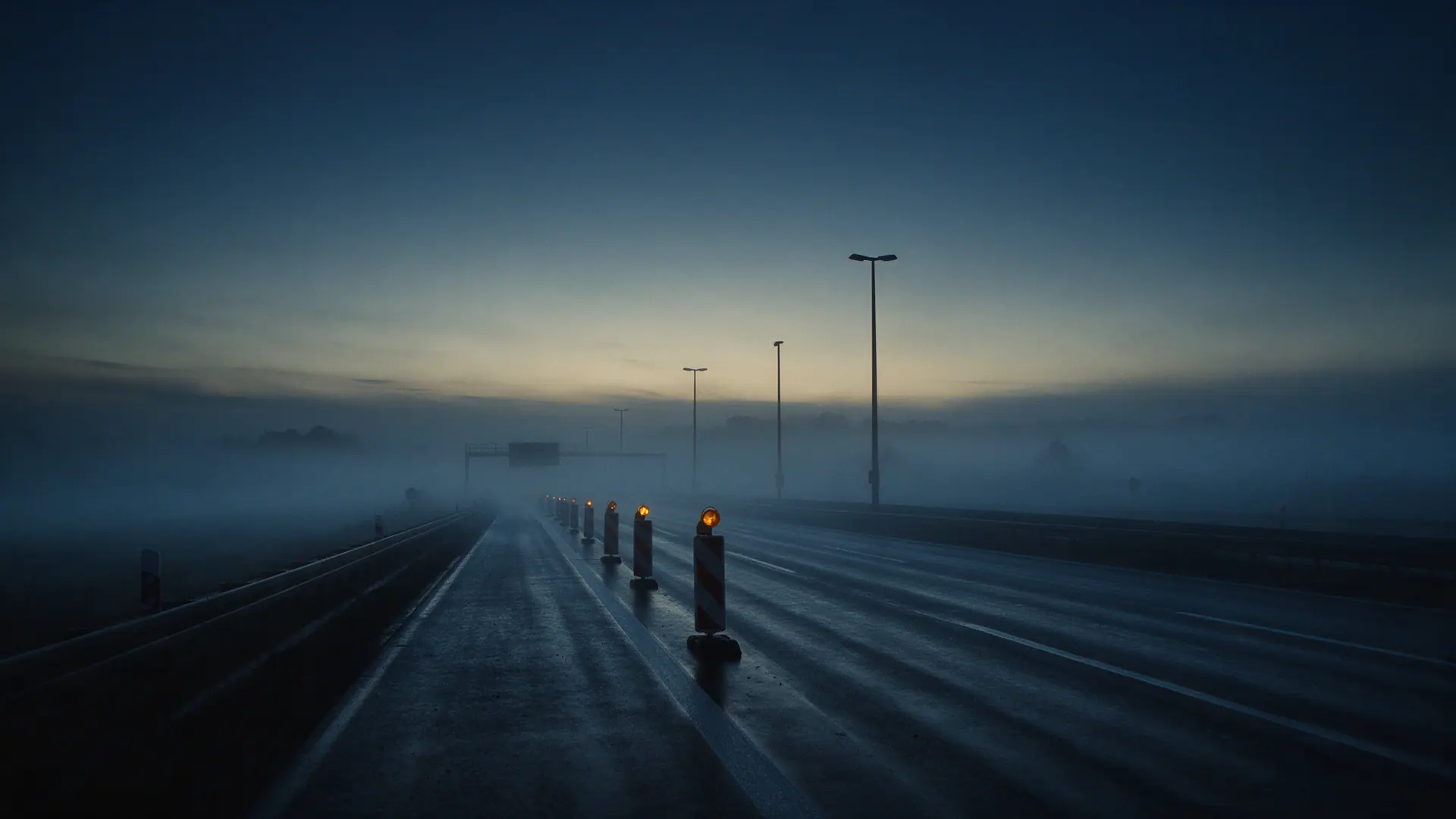 Leere Autobahn im Nebel kurz vor Sonnenaufgang, eine Leitbakenreihe mit brennenden Warnleuchten führt in den Dunst. Bildmotiv KI-generiert.