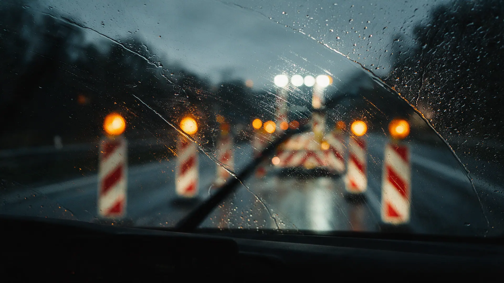 Blick durch eine regennasse Windschutzscheibe auf eine beleuchtete Arbeitsstelle, Leitbaken und Warnleuchten unscharf im Hintergrund. Bildmotiv KI-generiert.