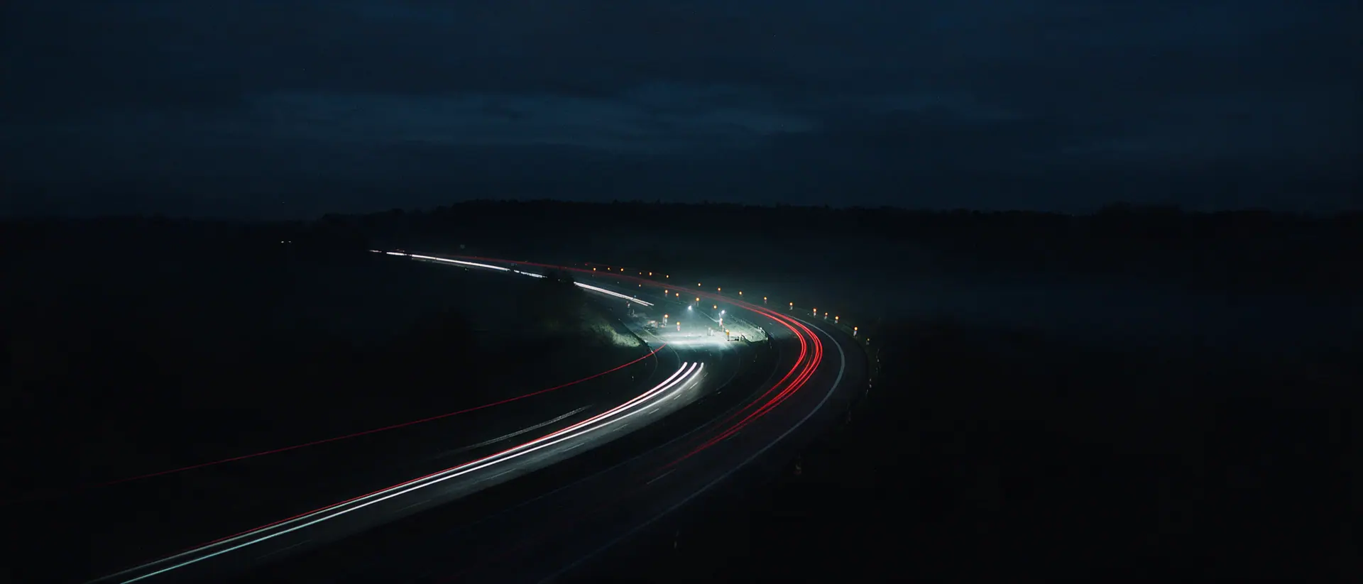 Autobahn bei Nacht aus erhöhter Position, Lichtspuren folgen der Kurve, ein beleuchteter Arbeitsstellenabschnitt liegt hell in der Dunkelheit. Bildmotiv KI-generiert.