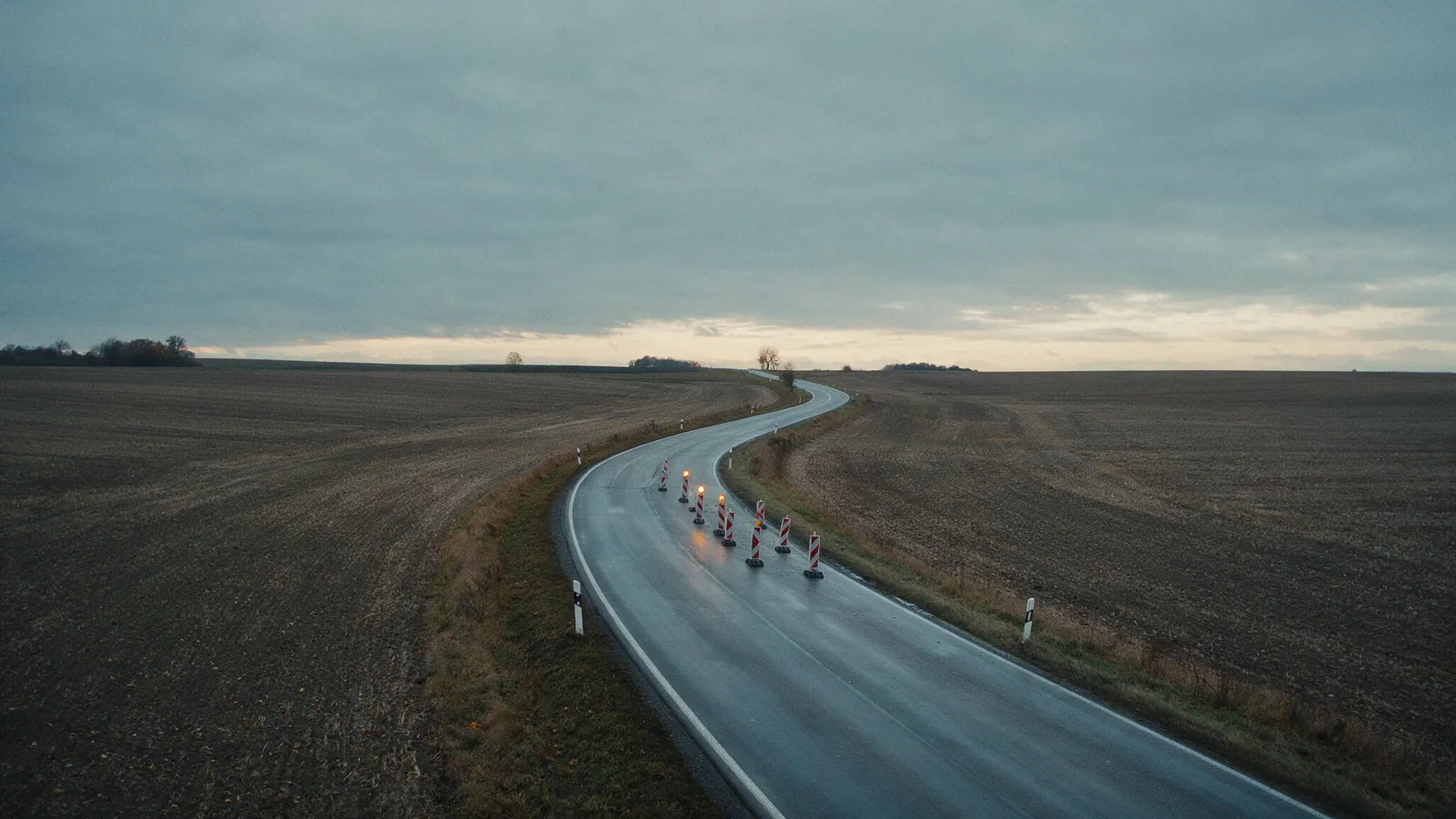 Weiter Blick über eine Landstraße in der Dämmerung, die zwischen kahlen Feldern verläuft; ein kurzer abgesperrter Abschnitt mit wenigen Leitbaken und zwei Warnleuchten liegt klein in der Landschaft. Bildmotiv KI-generiert.